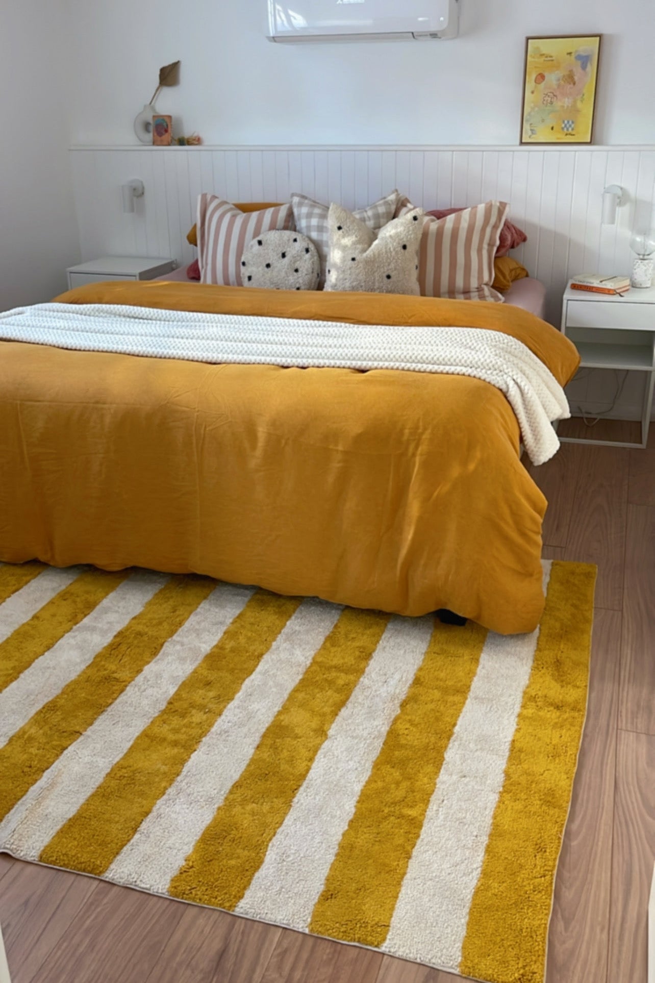 Bedroom with a bed covered in yellow and white bedding, striped rug, and air conditioner on the wall.